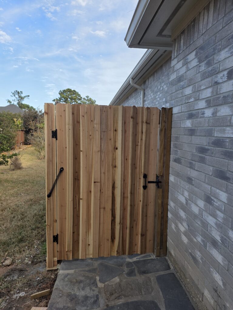 Vertical cedar wood gate with black hinges next to a brick wall.